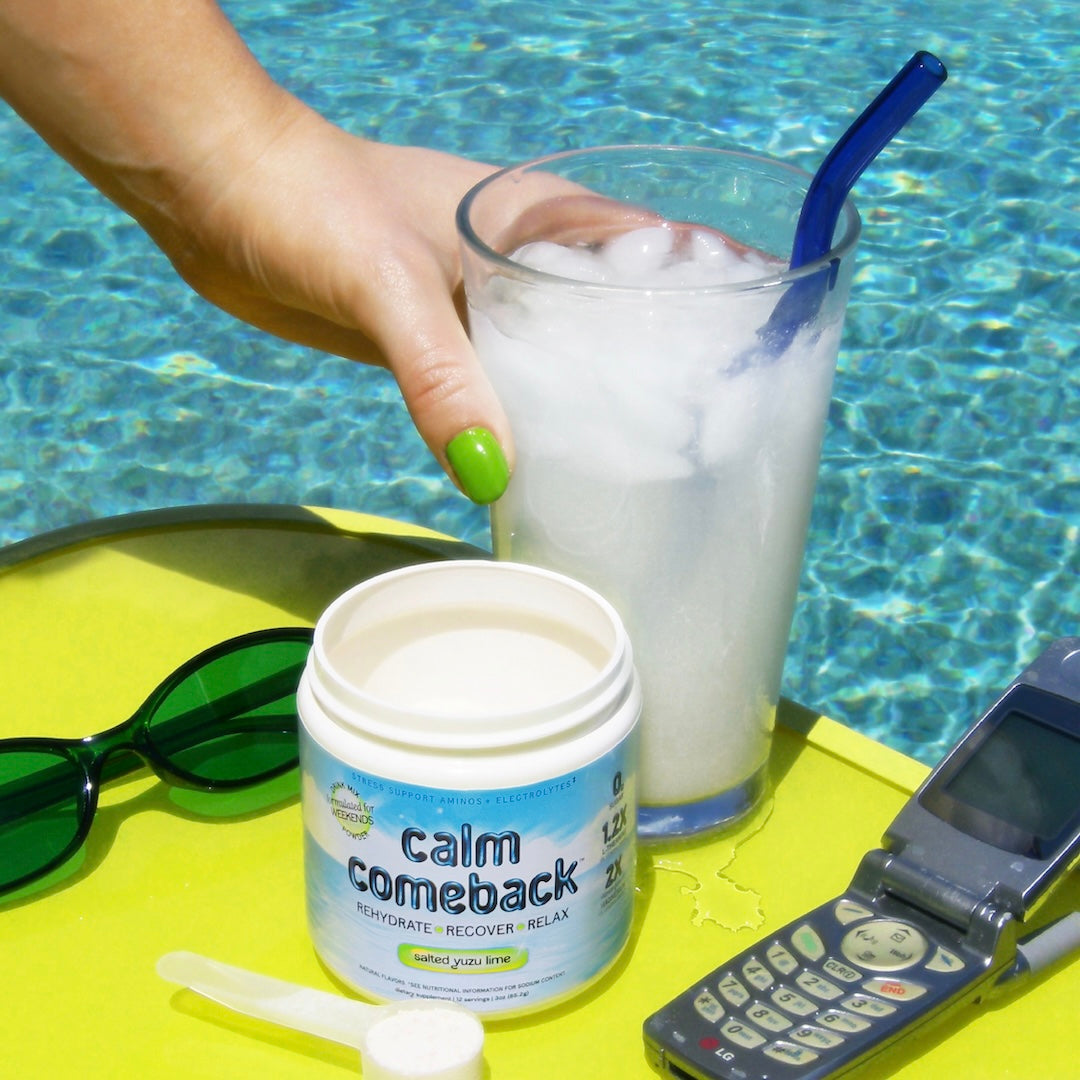A jar of Calm Comeback is shown on a yellow poolside table next to a glass of iced drink with a blue straw, sunglasses, and a flip phone, with a person's hand holding the drink.